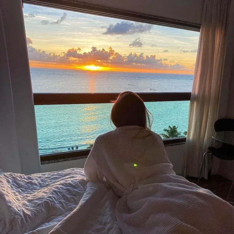 Uma mulher está deitada na cama olhando pela janela para o oceano.