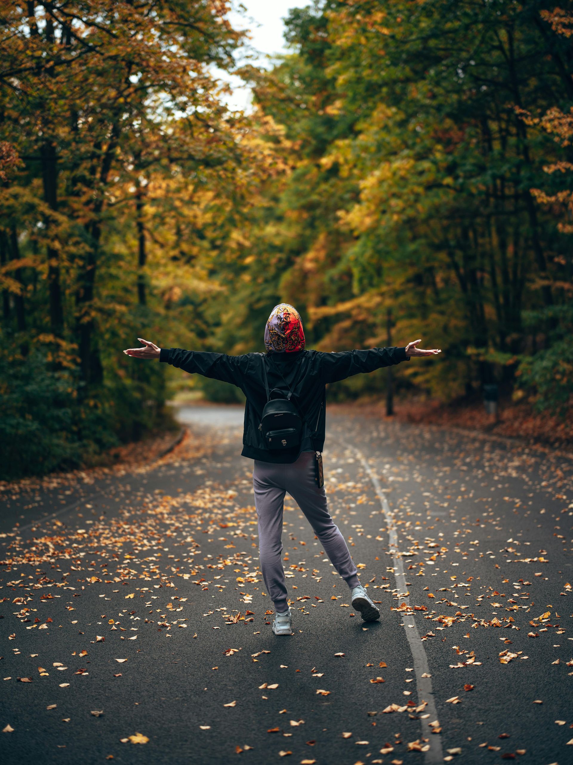 a person wearing a backpack looking down the road covered with leaves. the person has their arms out feeling relieved. 