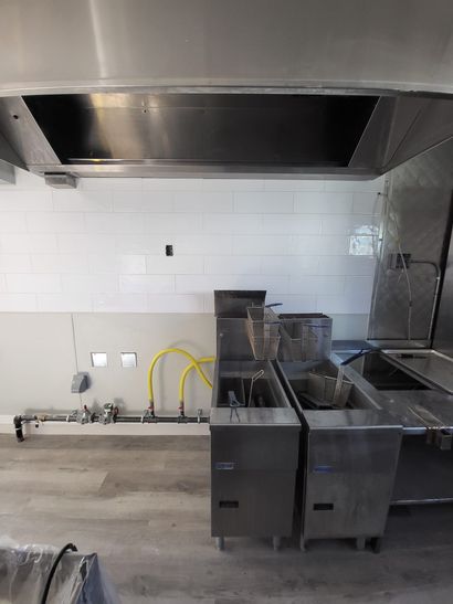 Empty commercial kitchen with stainless steel prep tables and large ventilation hood against a tiled wall.