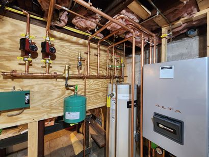 Basement utility area with plumbing pipes, pressure tank, and a large white furnace or boiler unit.