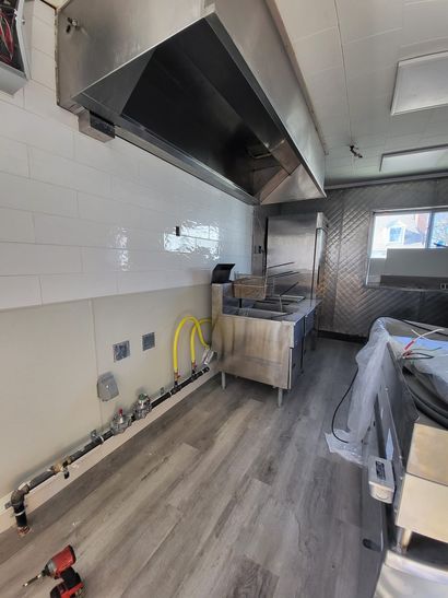 Empty commercial kitchen with stainless steel hood, white tile walls, and gray wood-look flooring.