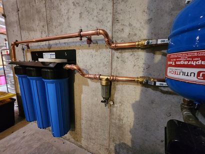 Water filtration setup with blue canisters, copper pipes, and a wall-mounted tank in a basement setting