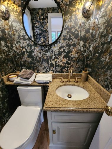 Small bathroom with floral wallpaper, white toilet, granite sink vanity, and oval mirror.