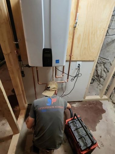 Technician kneels in a framed wall cavity beside a water heater, working with tools and wiring.