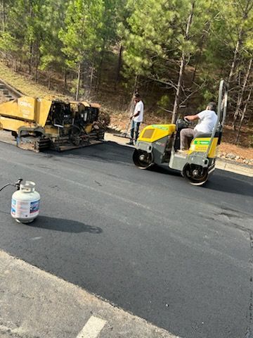 Road crew paving asphalt with a yellow machine beside a worker on a small cart, propane tank in foreground