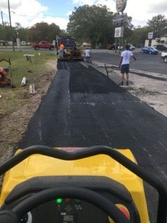 View from a yellow paving machine laying fresh black asphalt on a roadside, with workers nearby.