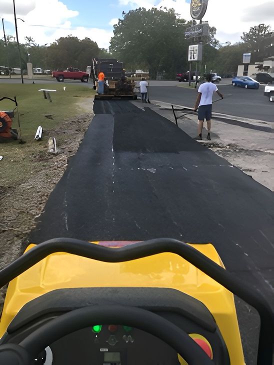 Paved walkway under construction beside a road, with workers paving asphalt and a yellow machine in the foreground.