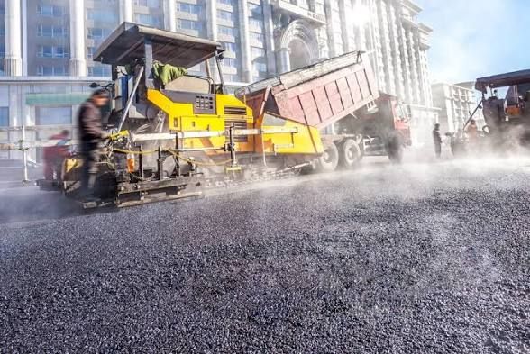 Road paver laying fresh asphalt on a city street beside a dump truck, with steam and dust rising