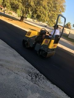 Small yellow road roller on a paved street near trees, viewed from behind.