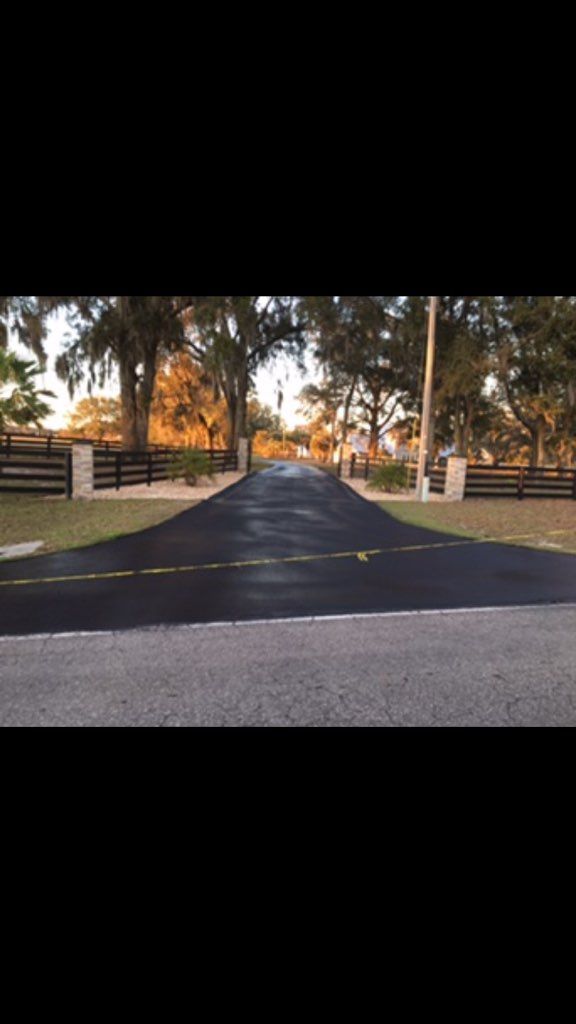 Roadway leading to a gated driveway at sunset, lined with trees and white fence posts.