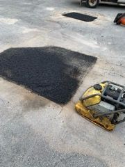 Black asphalt patch on a concrete road beside a yellow plate compactor and a parked vehicle