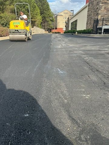 Road roller compacting fresh black asphalt on a driveway or road beside buildings and trees.