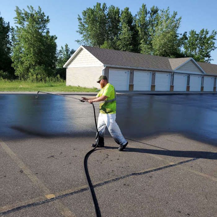 Worker pressure washing a large asphalt lot beside a row of white garages and trees.