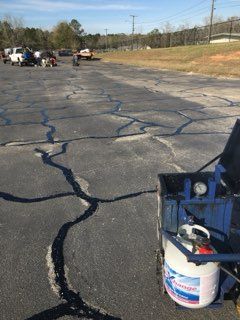 Cracked, damaged parking lot with a white paint sprayer in the foreground and work vehicles in the distance