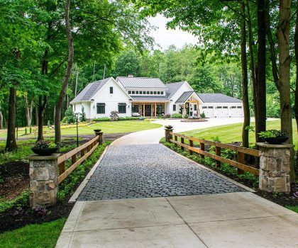White house with black roof, wooden accents, and a stone and wood fence, with a paved driveway in a wooded setting.