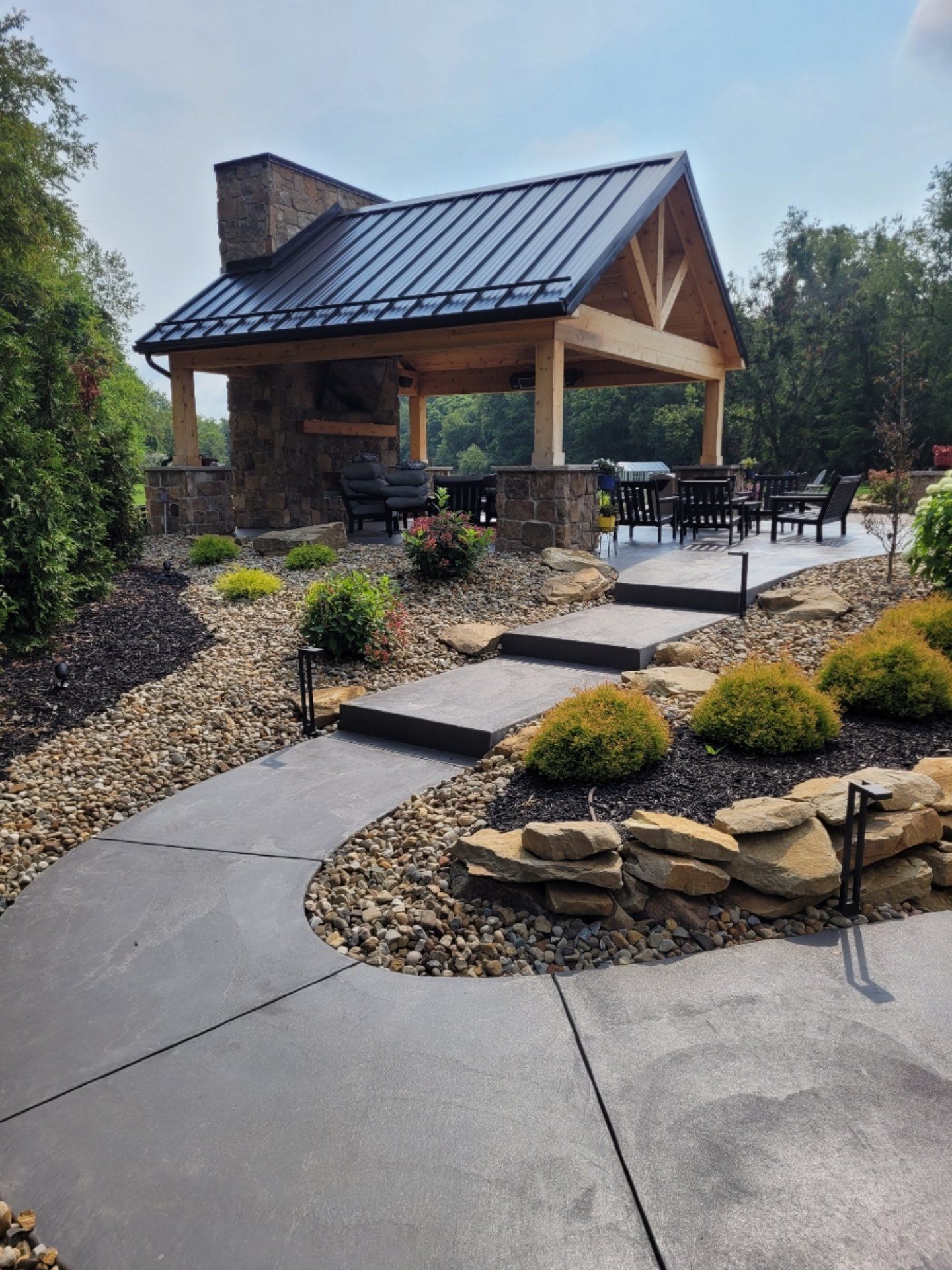 Stone and wood outdoor pavilion with fireplace, black roof, and patio. Pathway leads to seating area.