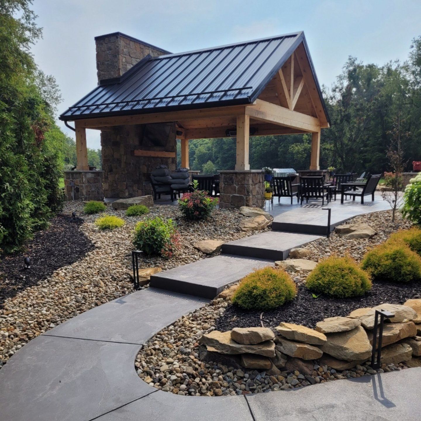 Stone and wood pavilion with a fireplace and black metal roof. Steps lead up to a patio.