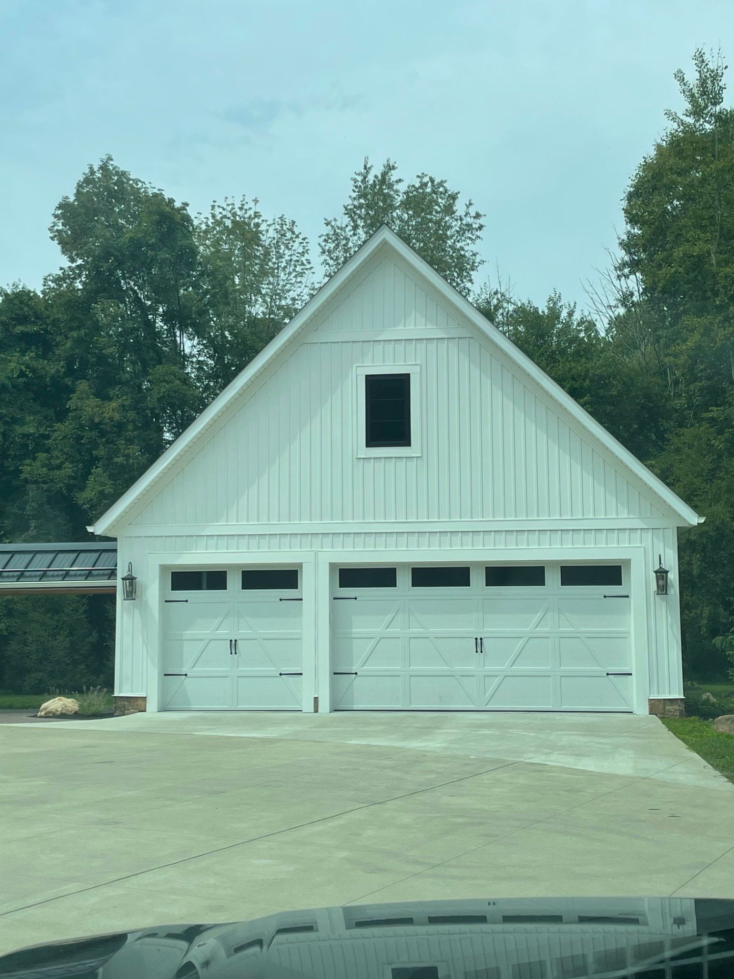 White barn-style garage with two bays, small window, and light fixtures. Concrete driveway. Trees in background.
