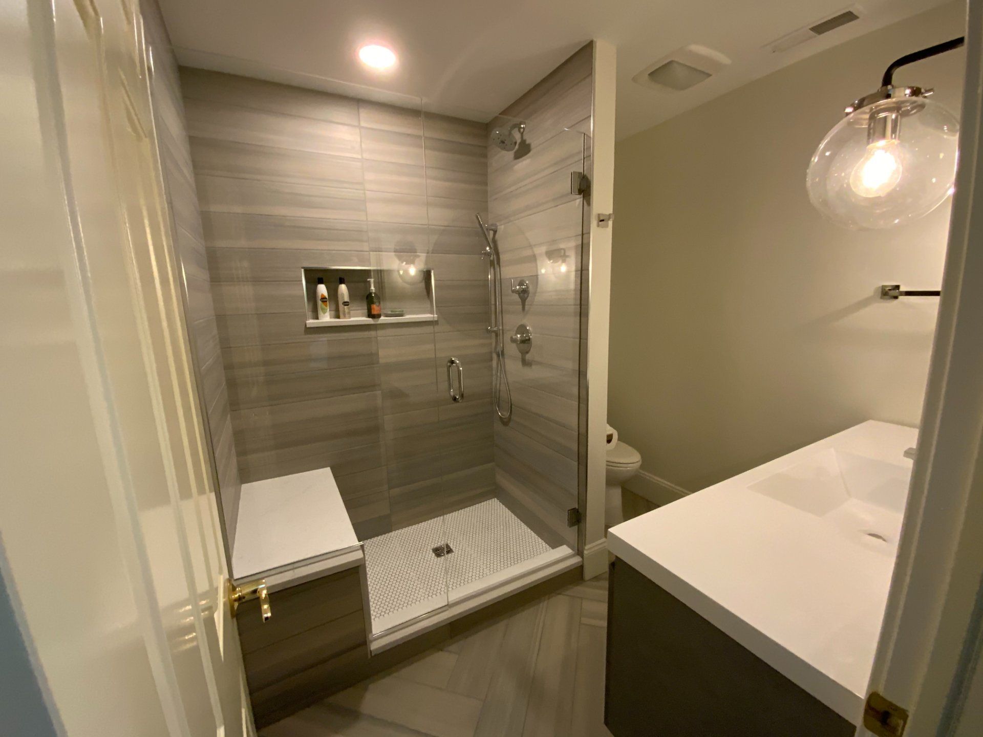 Modern bathroom with tiled shower, bench, and vanity. Neutral colors, glass door, and overhead lighting.