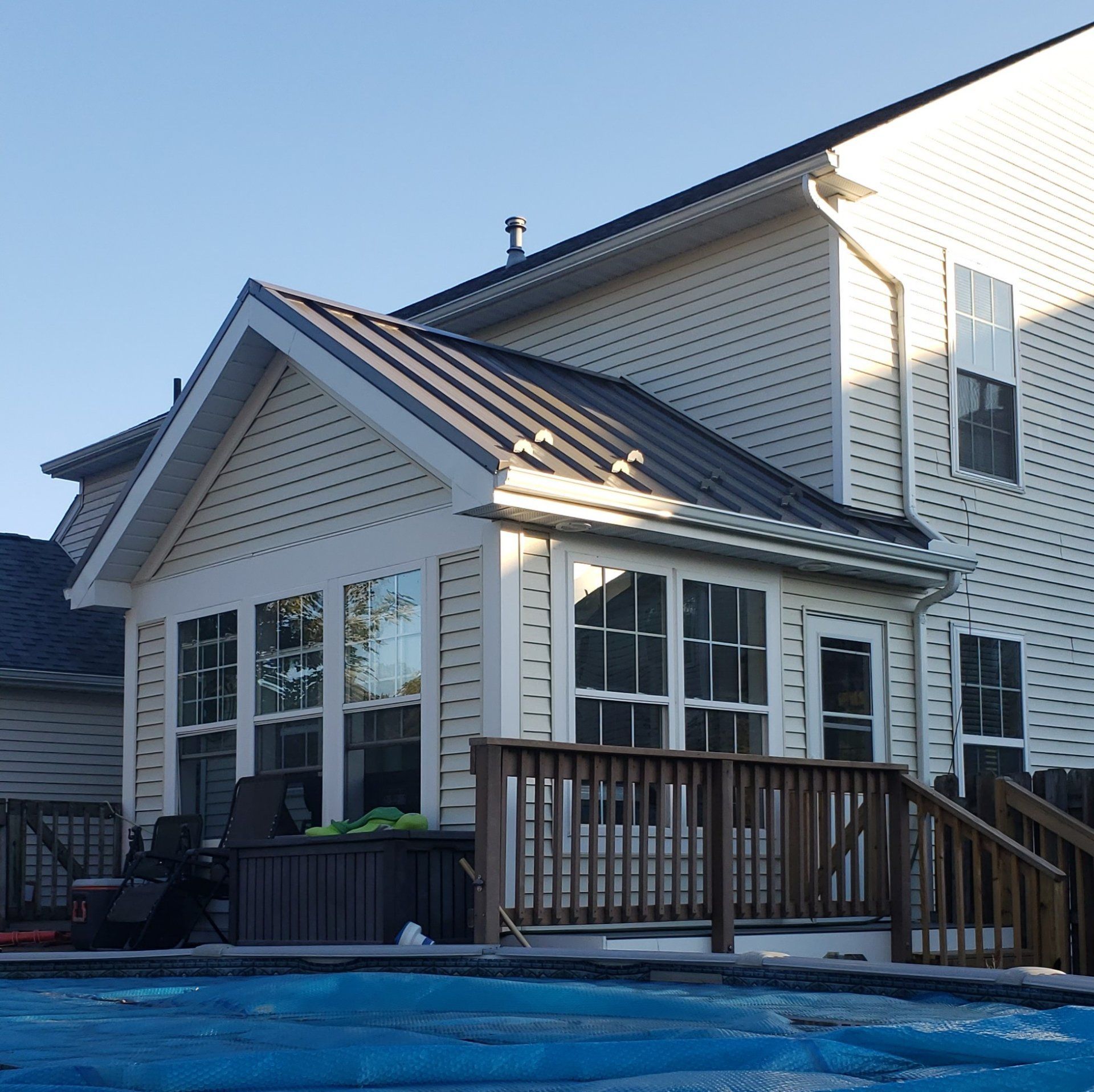 Sunroom with dark metal roof, adjacent to a house with white siding, next to a deck and pool.