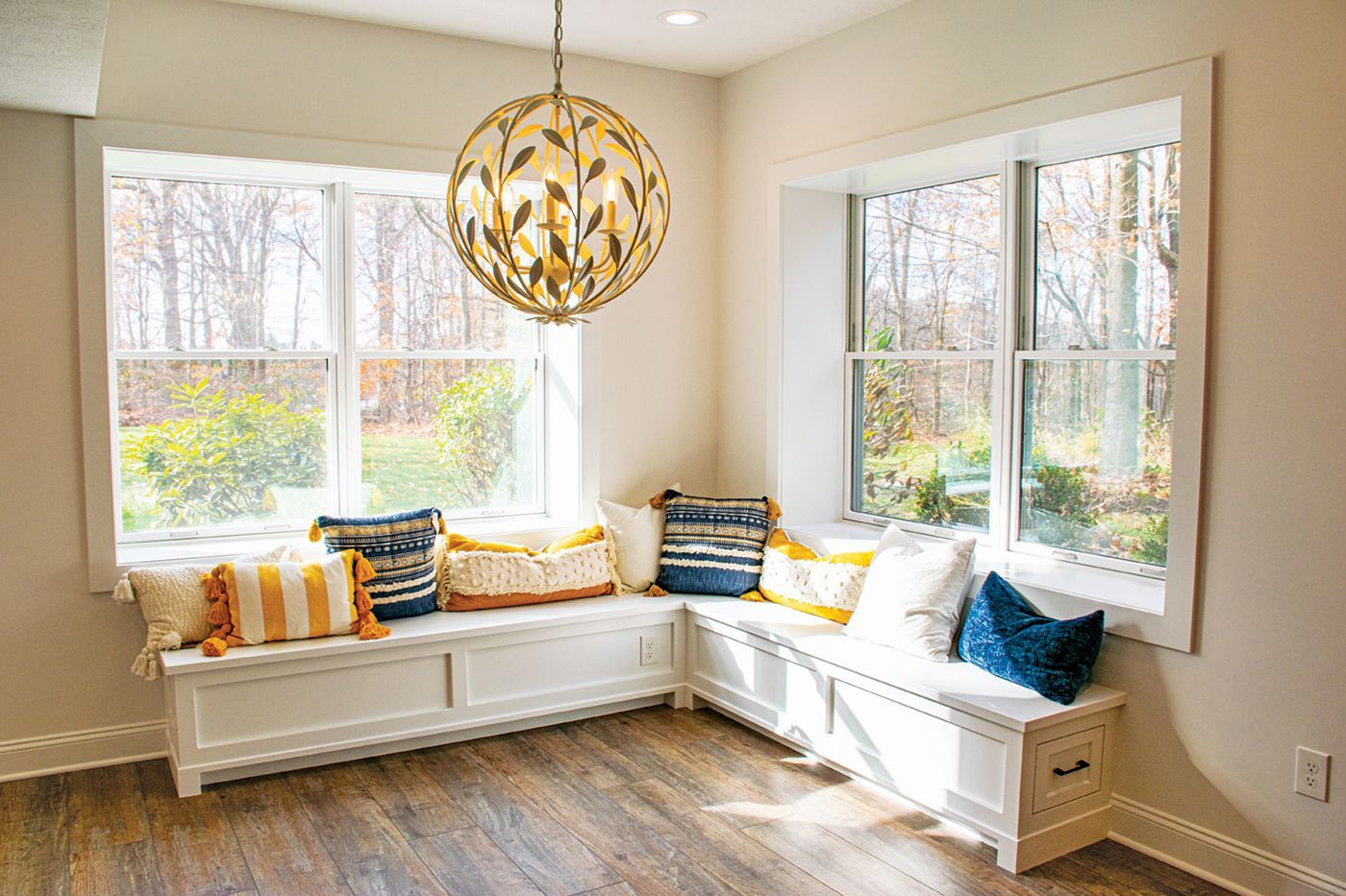 Corner window seat with storage, white trim, wood floor, colorful pillows, and gold light fixture.