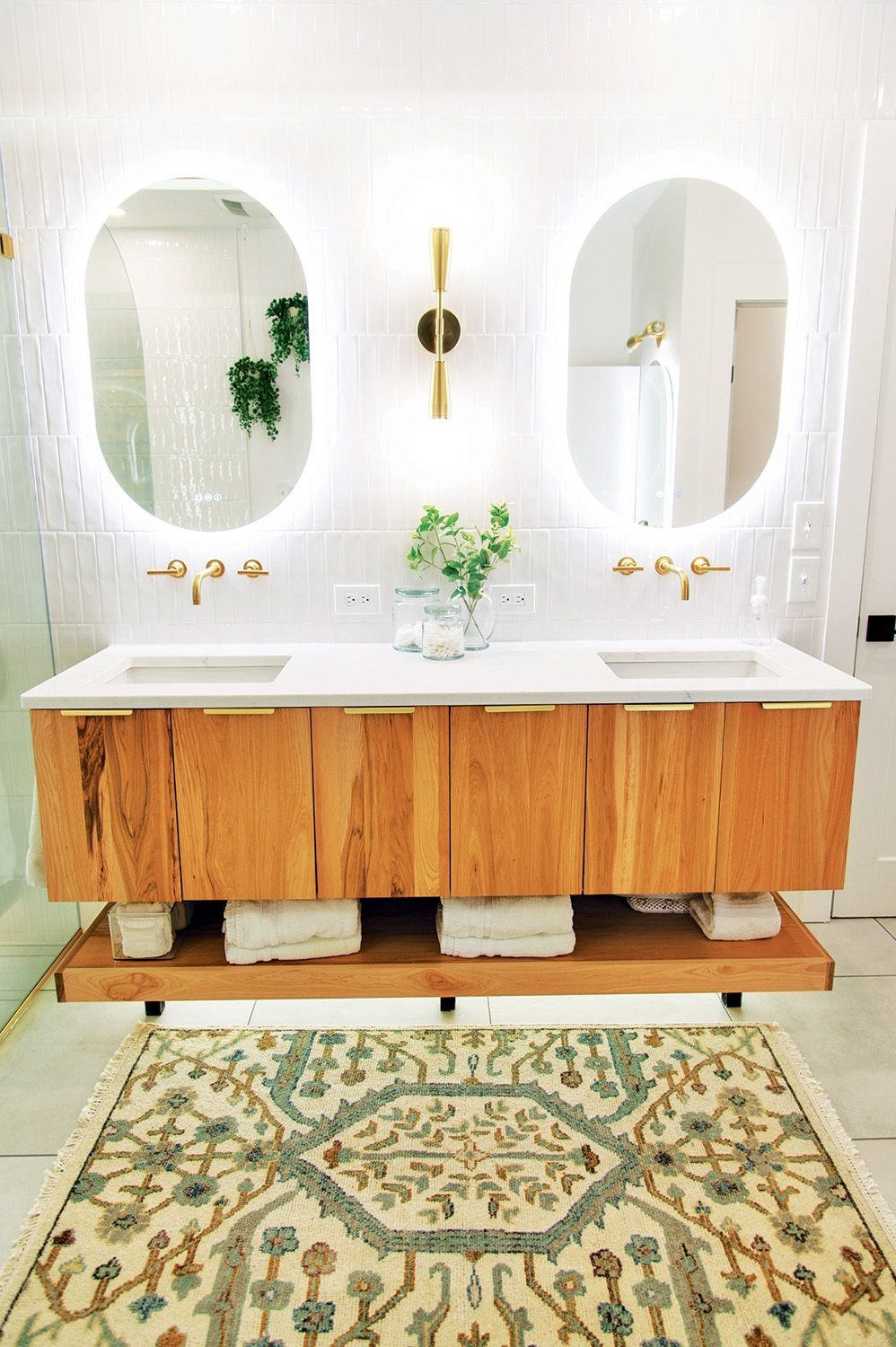 Modern bathroom with floating wooden vanity, oval mirrors with lights, and patterned rug.