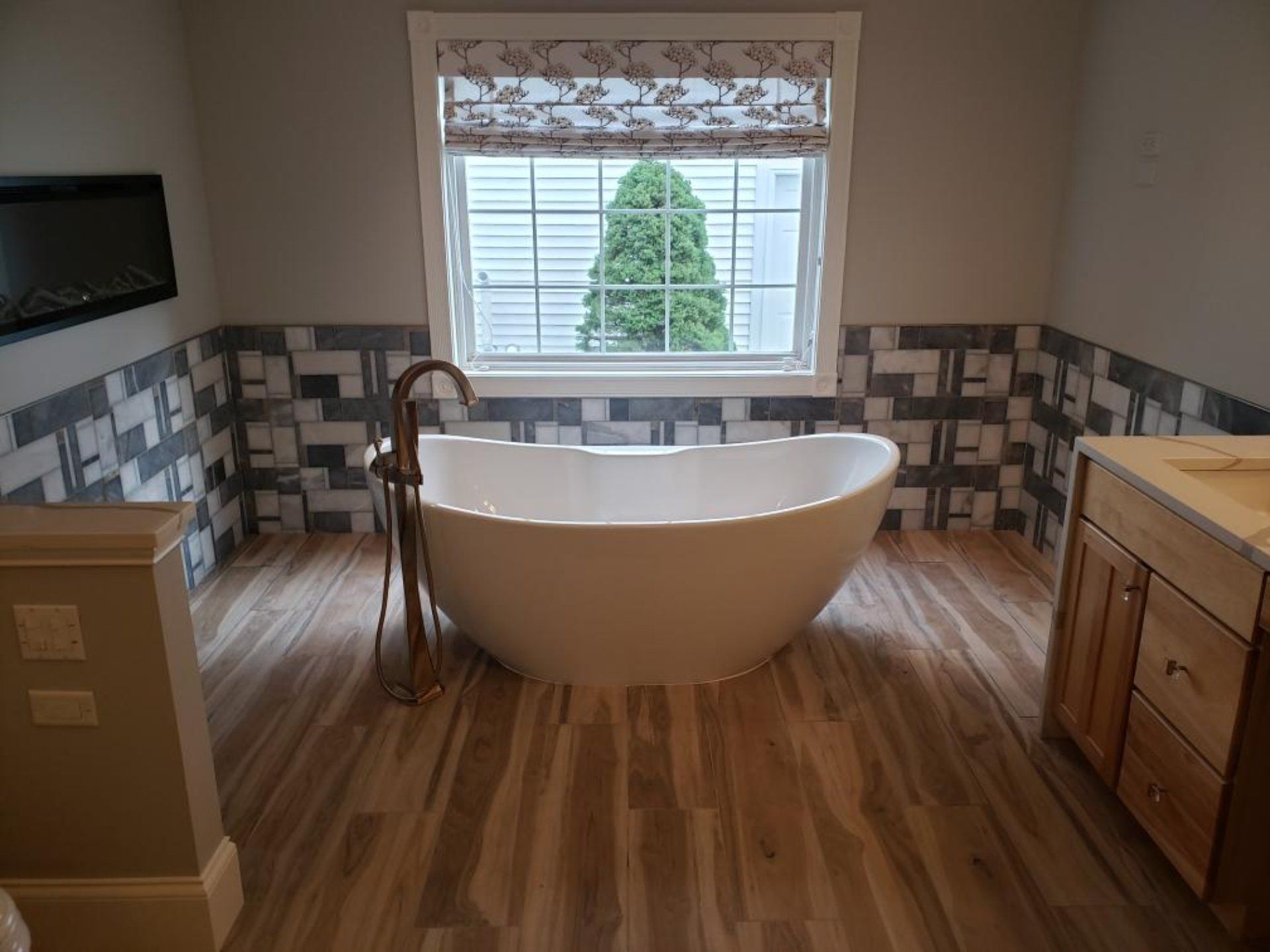 Bathroom with oval tub, wood-look floor, patterned tile wall, window with Roman shade, and a vanity.