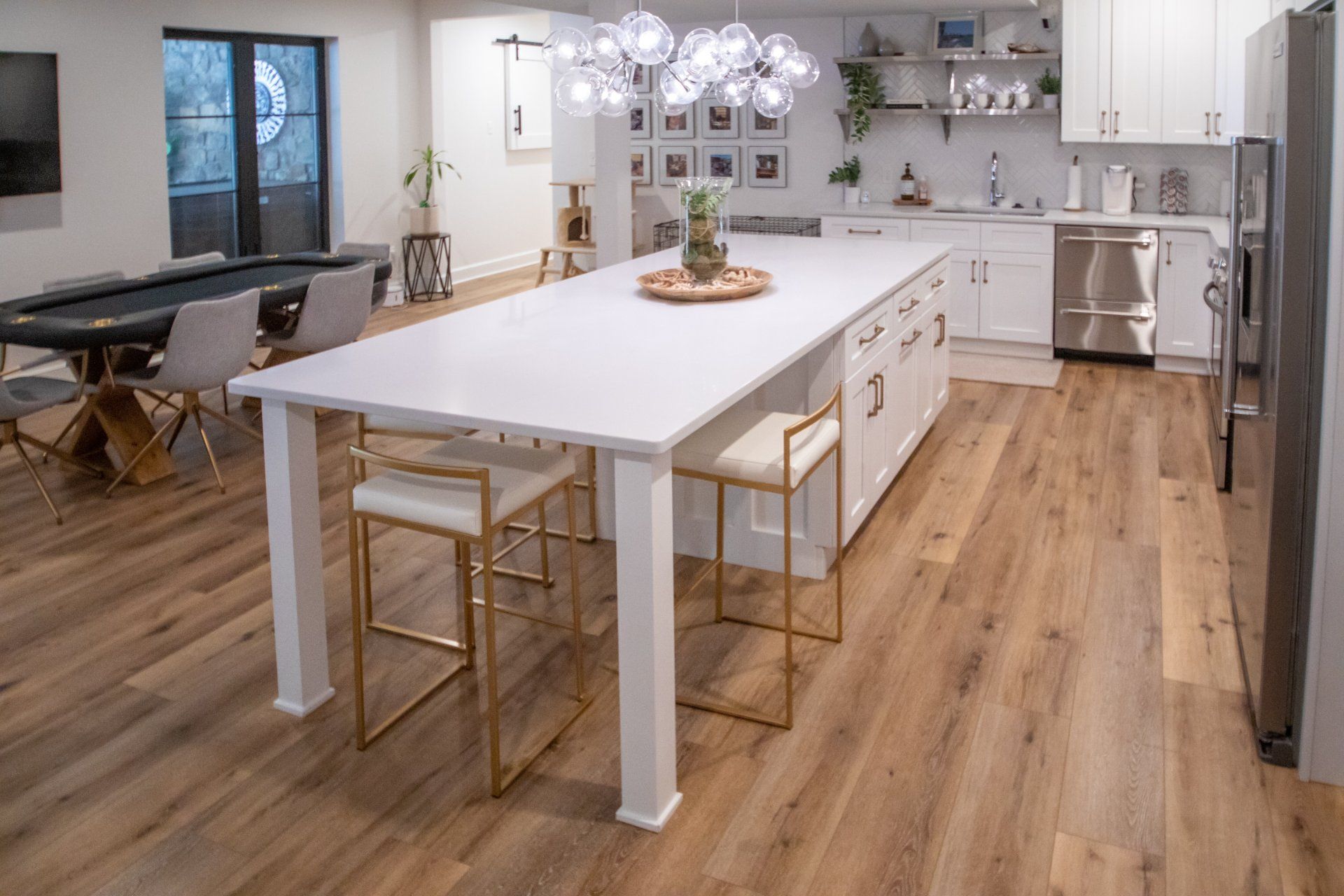 Modern kitchen with large white island, gold stools, and a poker table. Light wood flooring.
