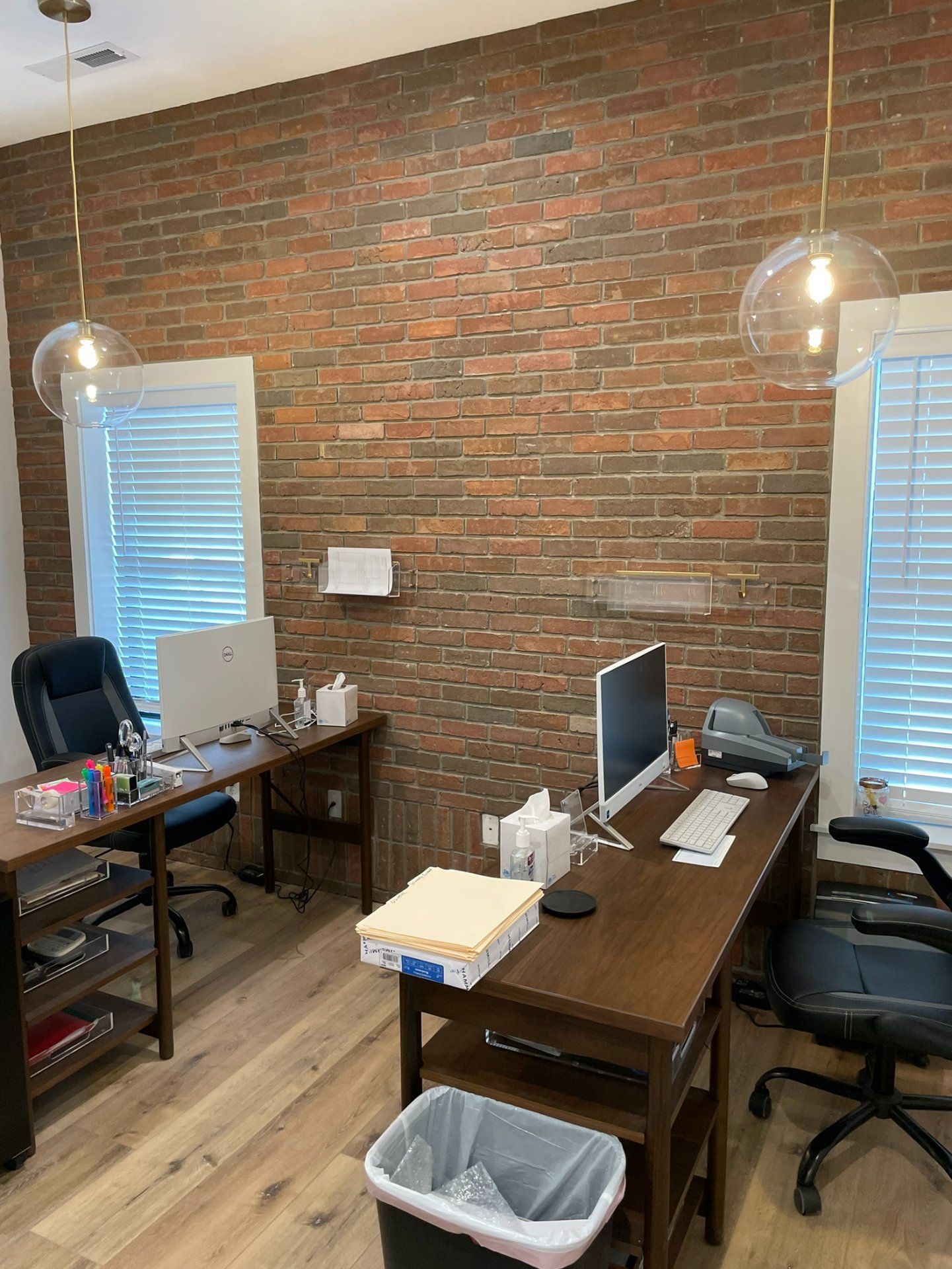 Office space with two desks against a brick wall, two pendant lights, two windows with blinds, and hardwood floors.