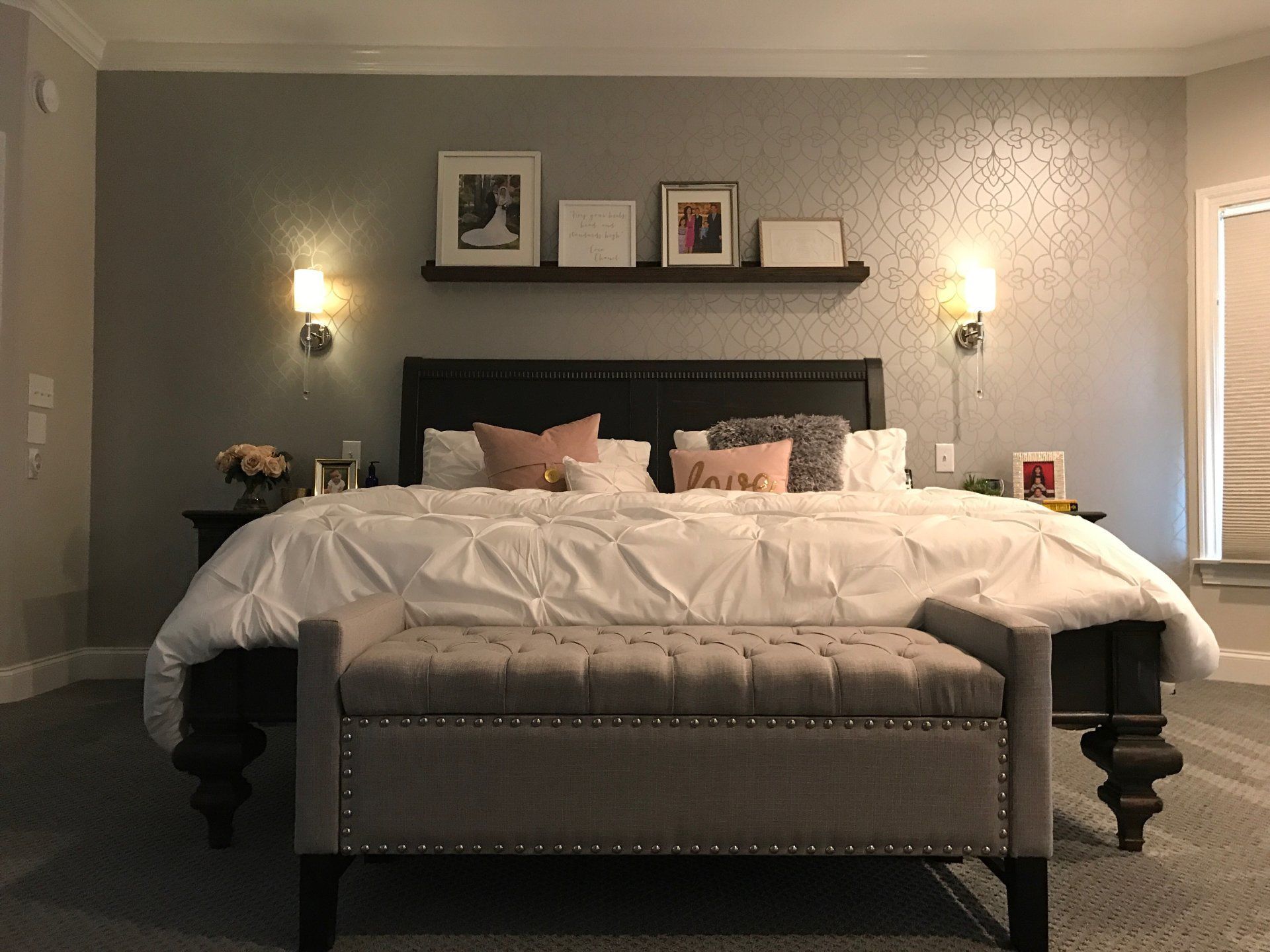 Bedroom with gray patterned wallpaper, dark bed frame, white bedding, and a gray tufted bench.