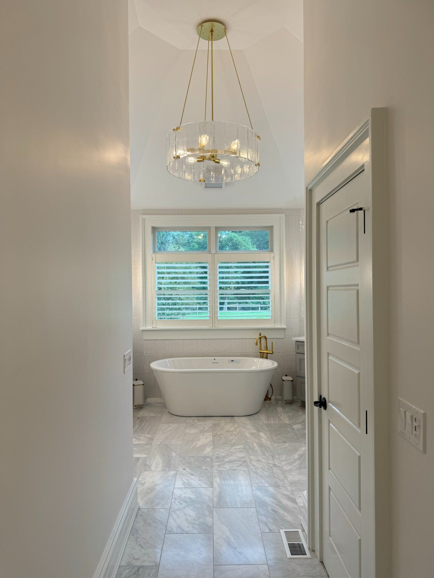 Narrow bathroom hallway with freestanding tub under a window, gold chandelier. White walls, grey tiled floor.