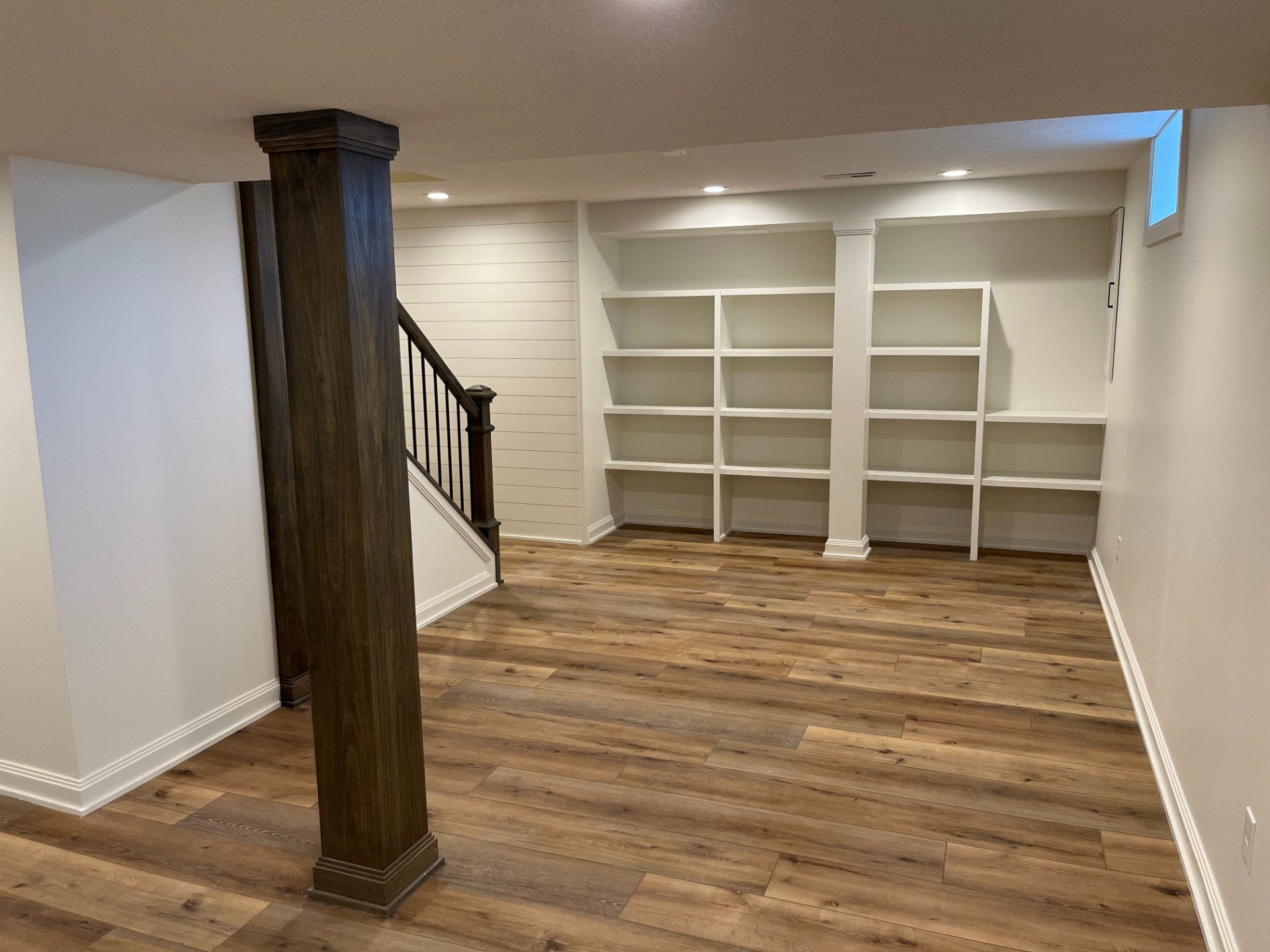 Basement with wood-look flooring, built-in white shelves, dark brown support column, and a staircase.