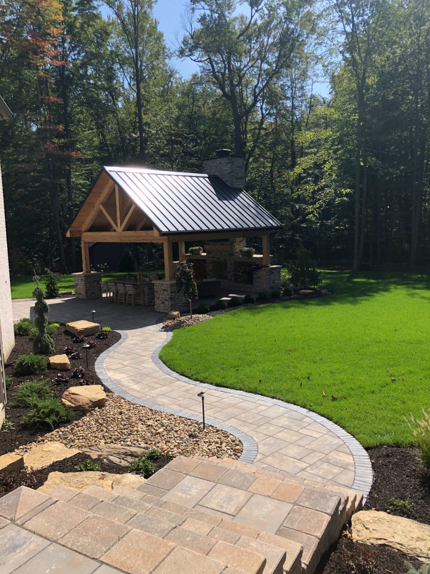 Stone path leads to a wooden pavilion with a metal roof. Backyard with green grass and trees.
