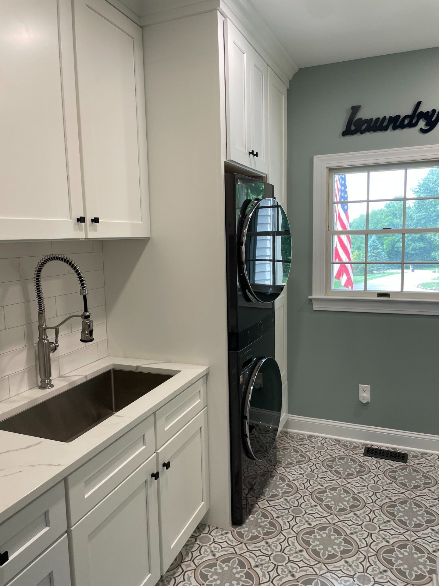 Laundry room with white cabinets, silver sink, stacked washer and dryer, patterned floor, and window.