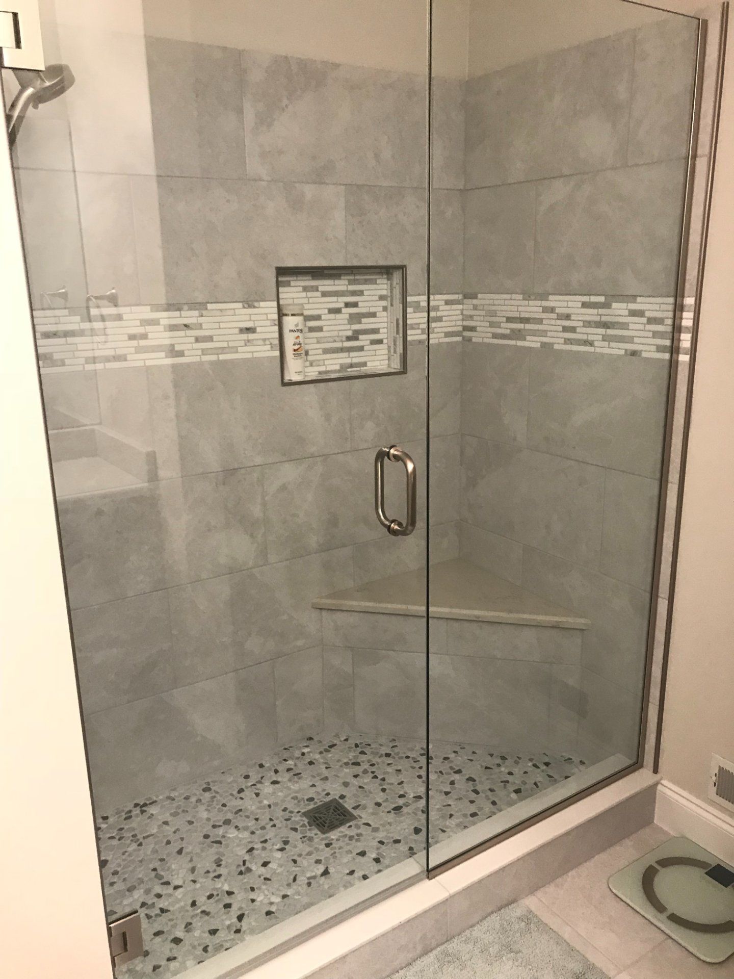 Shower with gray tile walls, pebble floor, glass door, and recessed shelf.