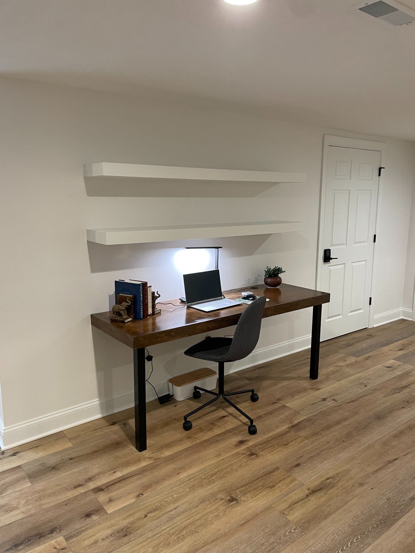Home office with desk, chair, laptop, shelves, and a closed door. Light wood floor and white walls.