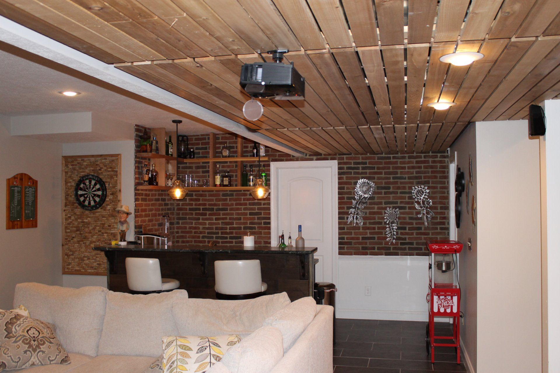 Basement entertainment area with a bar, brick accent wall, and wood-slat ceiling, including a projector.