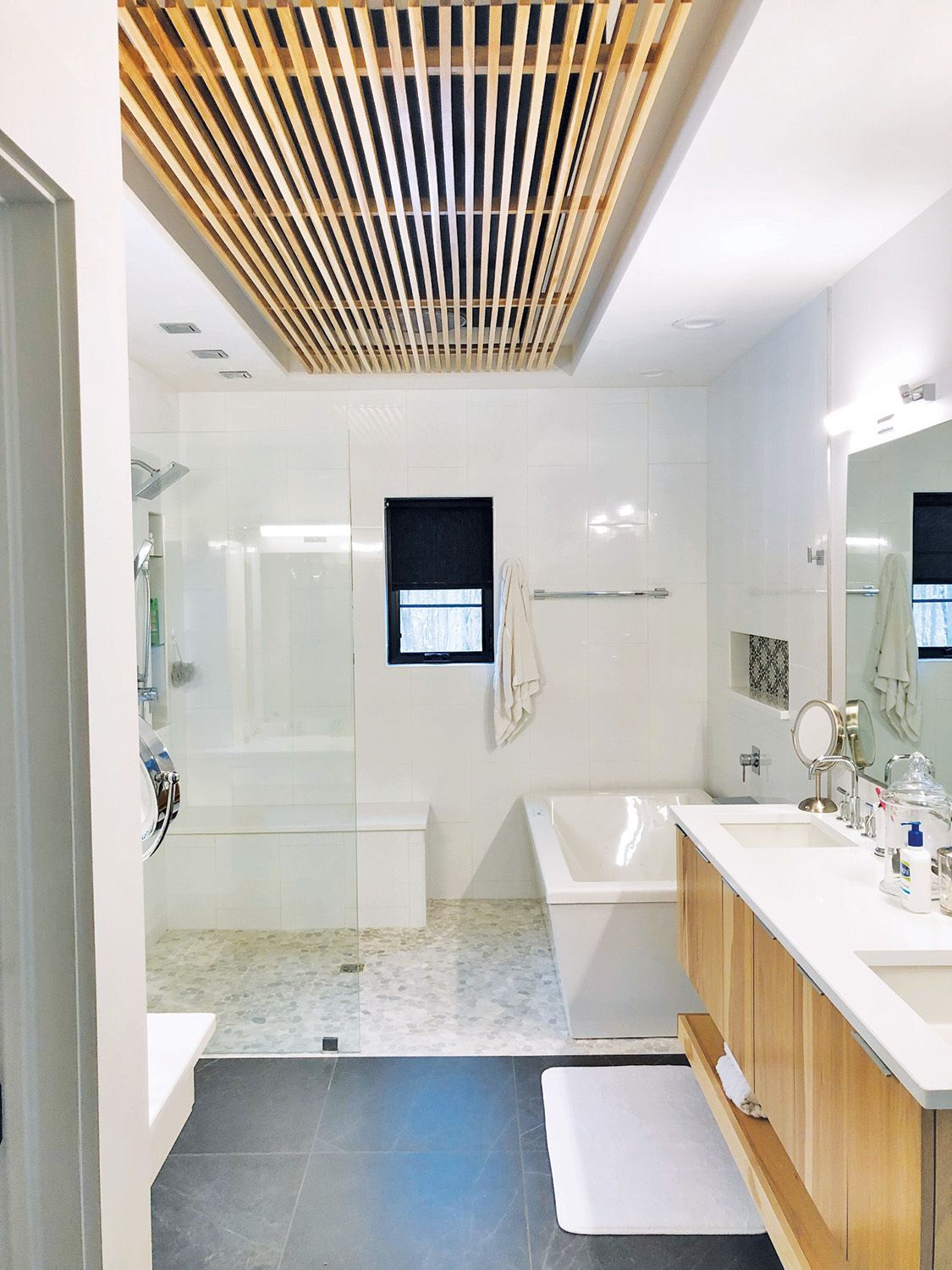 Modern white bathroom with wood-slatted ceiling, shower, bathtub, double vanity, and small window.