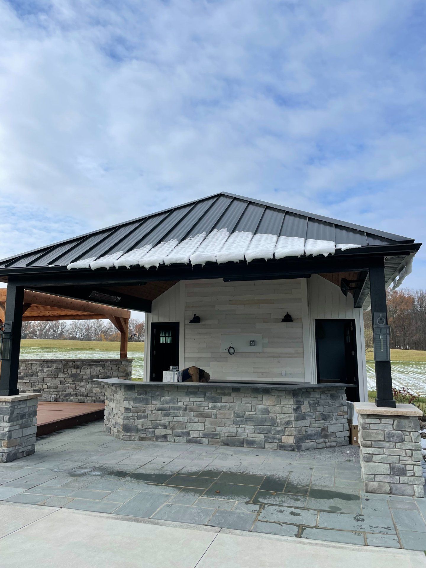 Outdoor pavilion with stone bar, black roof, and snow. Dark doors and fixtures, blue sky.