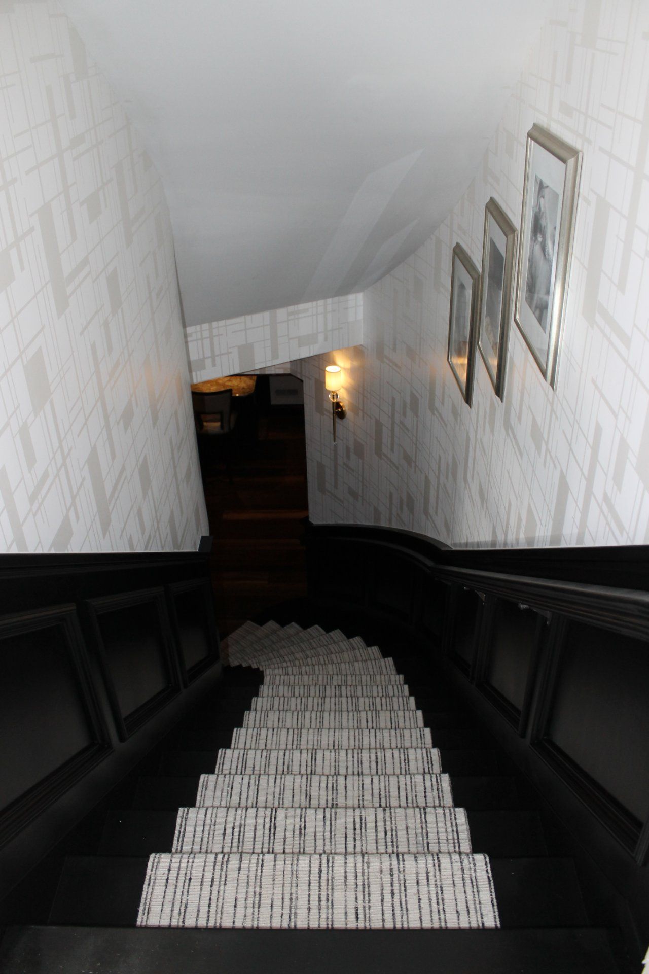 View down a staircase. Black treads with patterned runner, white patterned wallpaper, framed art on the wall.