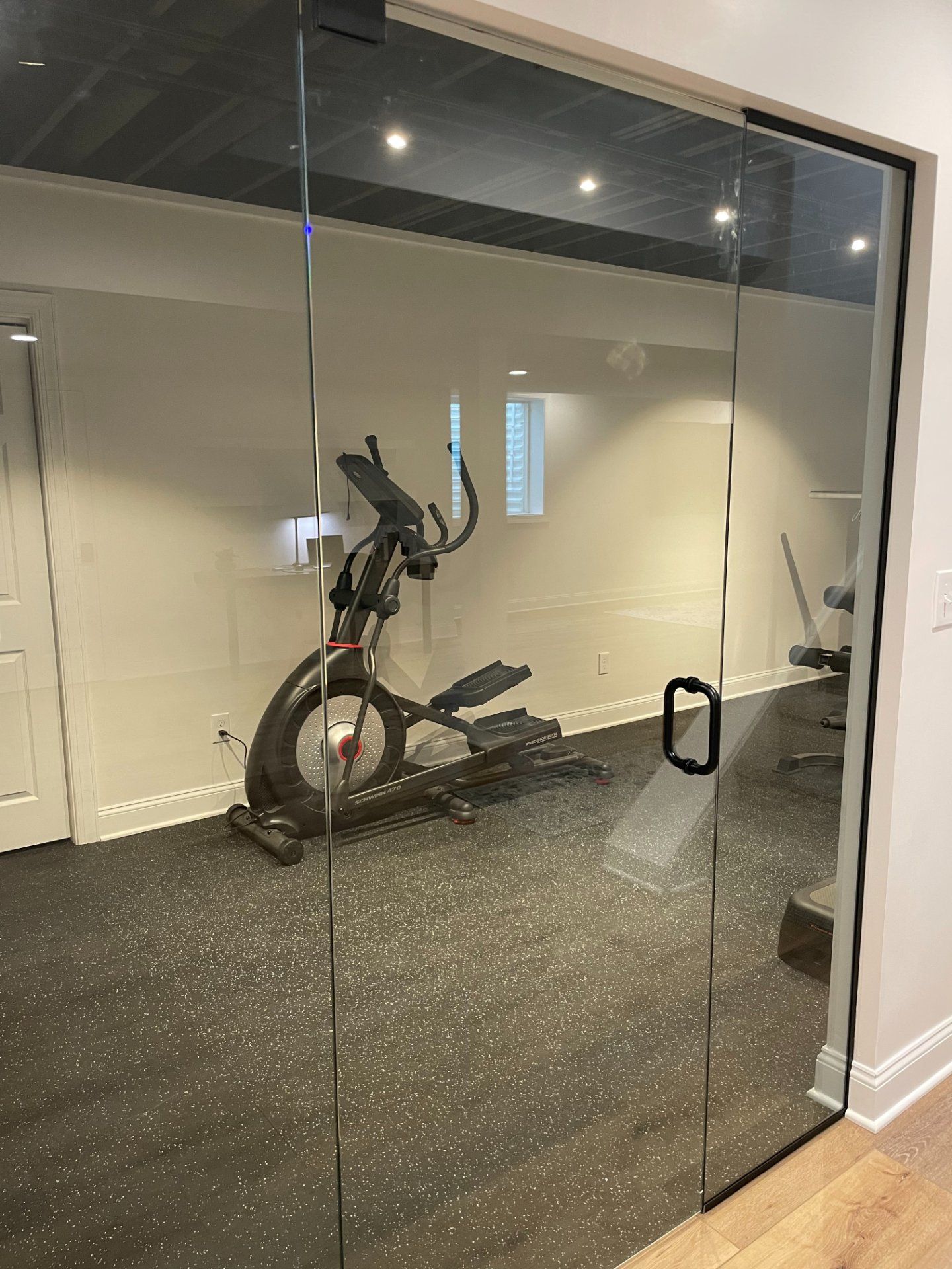 Glass-walled home gym with elliptical machine on speckled black flooring; door handle visible.