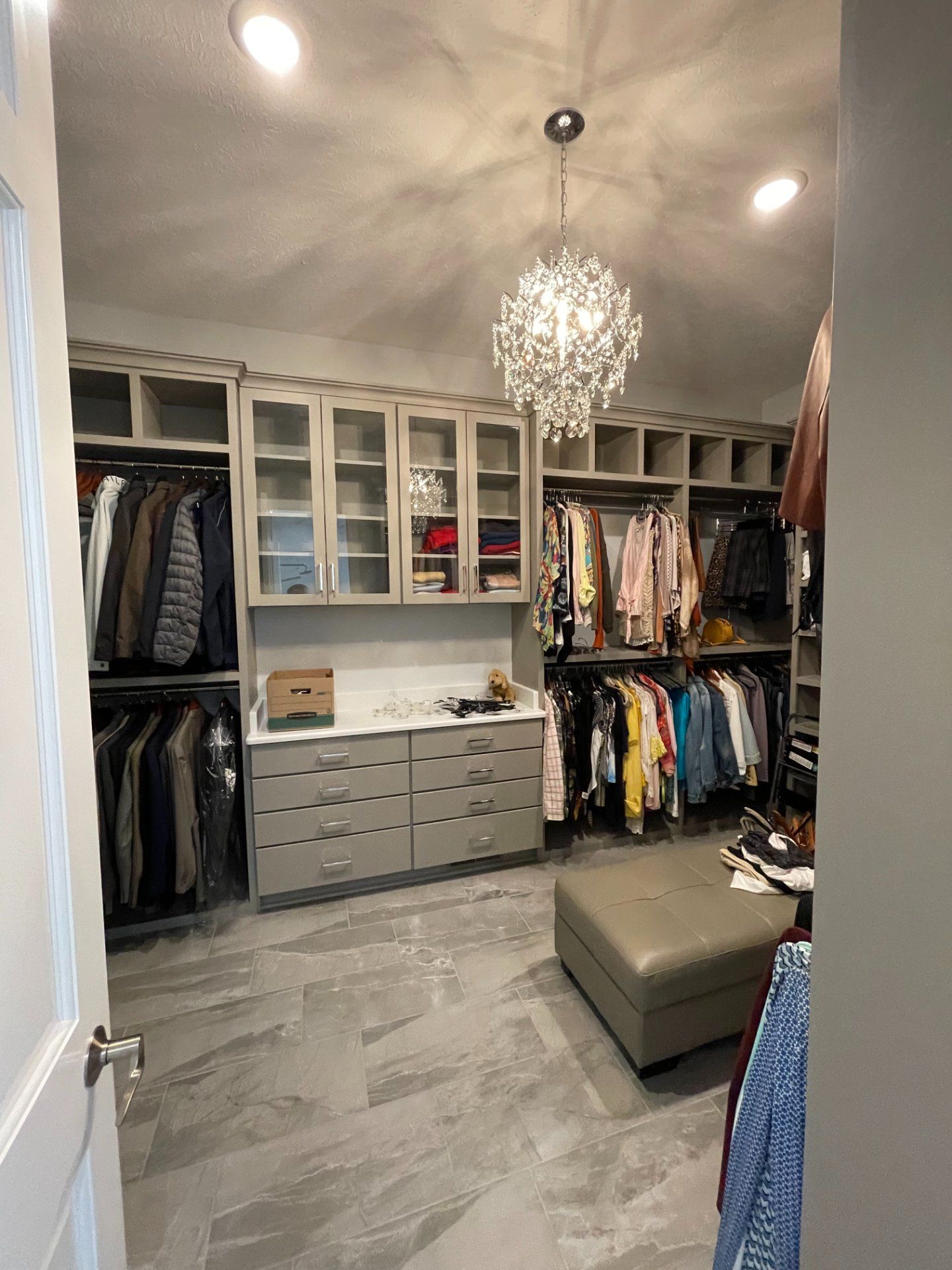Walk-in closet with gray cabinets, hanging clothes, drawers, and a chandelier; neutral tones.