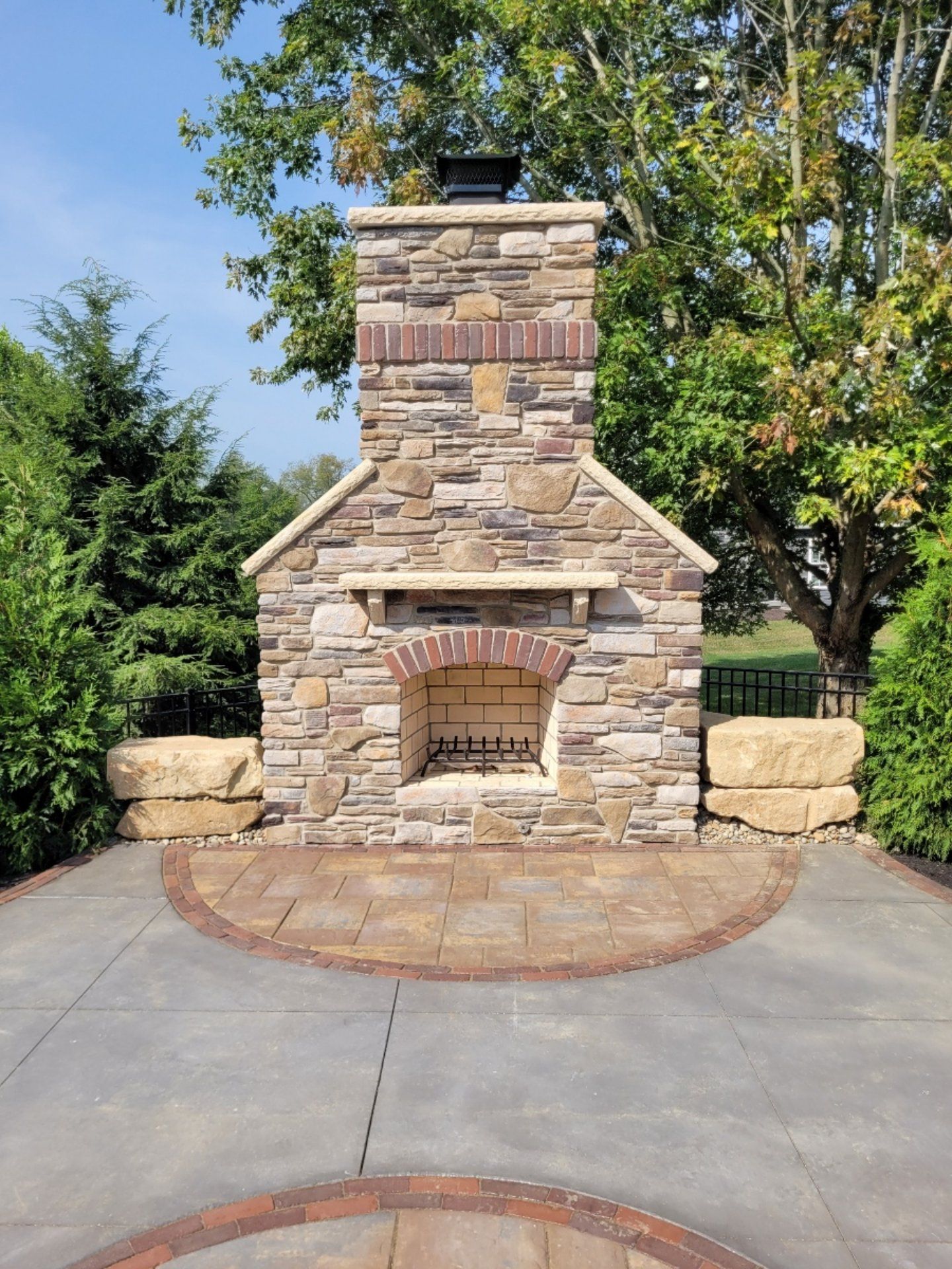 Stone outdoor fireplace with brick accents, surrounded by greenery and a paved patio.