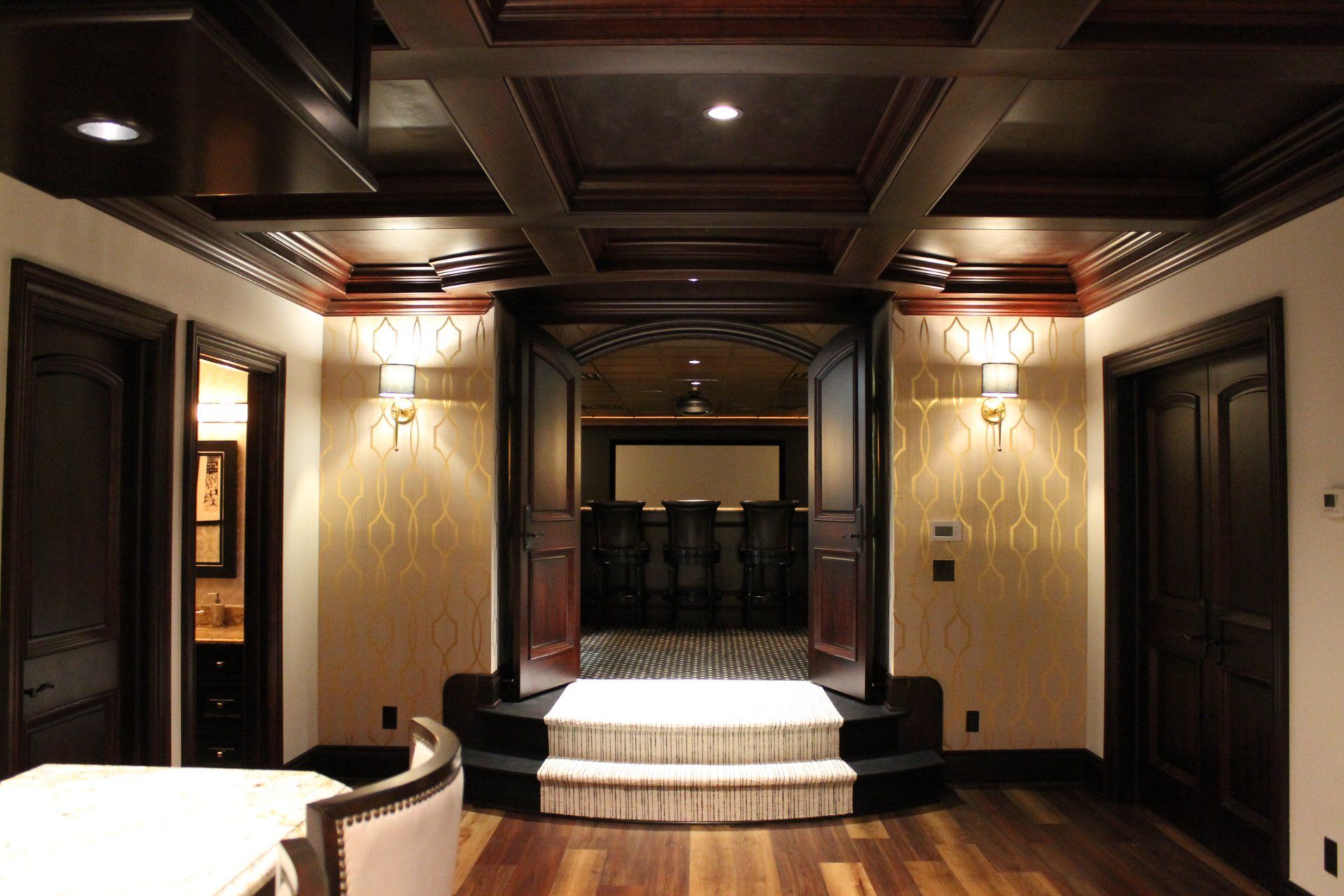Elegant, dark wood-paneled room with a multi-level floor, ornate ceiling, and doors leading to a bar area.