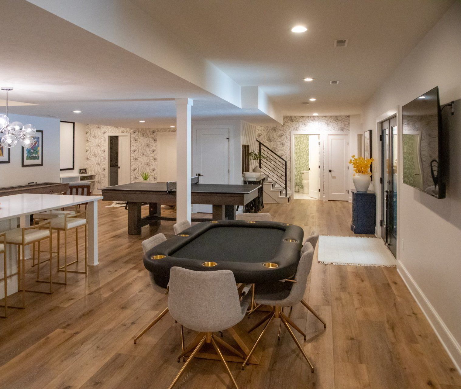 Spacious finished basement with a poker table, game tables, bar seating, and a TV. Wooden floors and white walls.