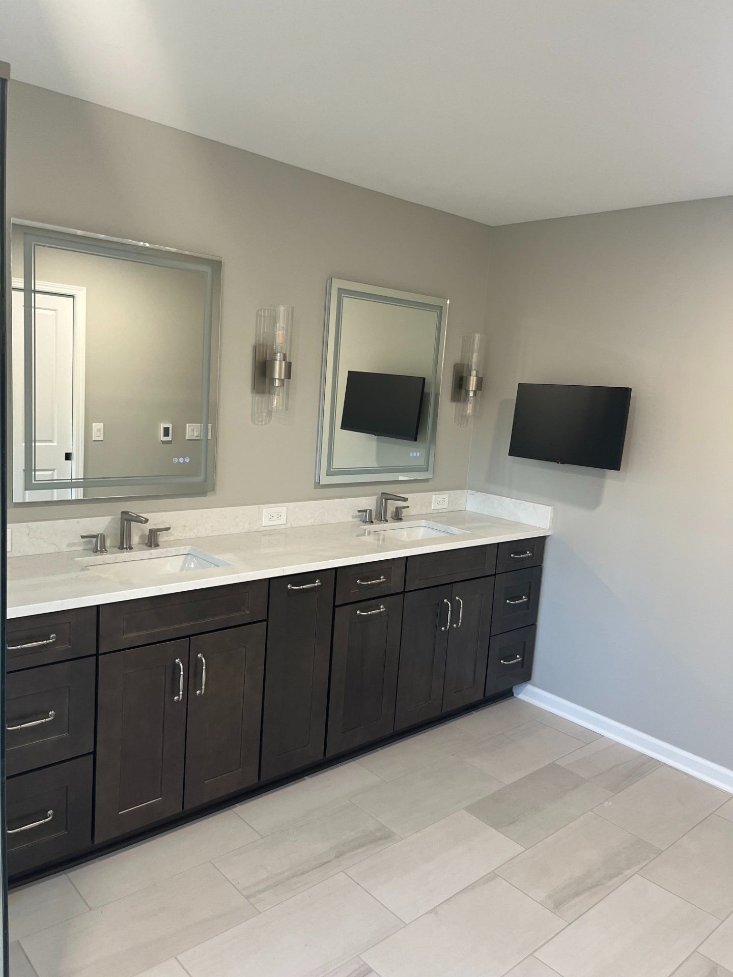 Bathroom with double sinks, two mirrors, and a wall-mounted TV. Brown cabinets, light countertops, and tan walls.
