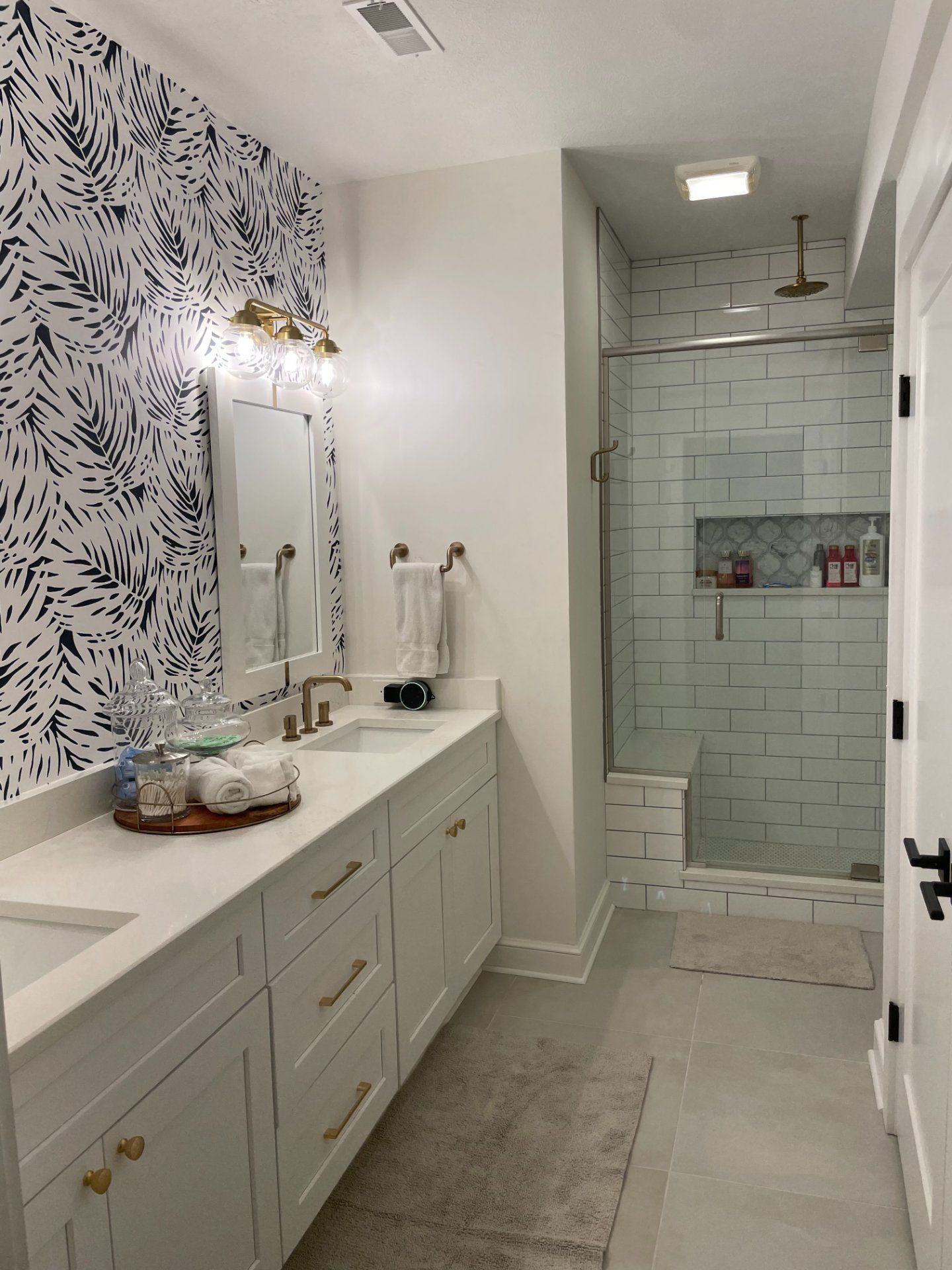 Bathroom with white cabinets, patterned wallpaper, a glass shower, and gold accents.