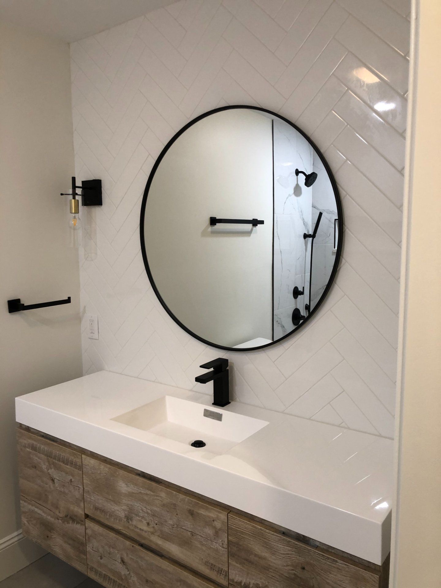 Modern bathroom with white herringbone tile, round black mirror, and wood-tone vanity.