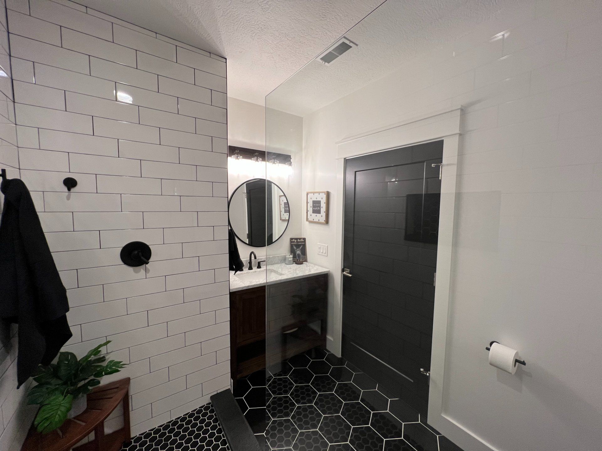 Bathroom with black and white tile, dark door, round mirror, and a small plant.