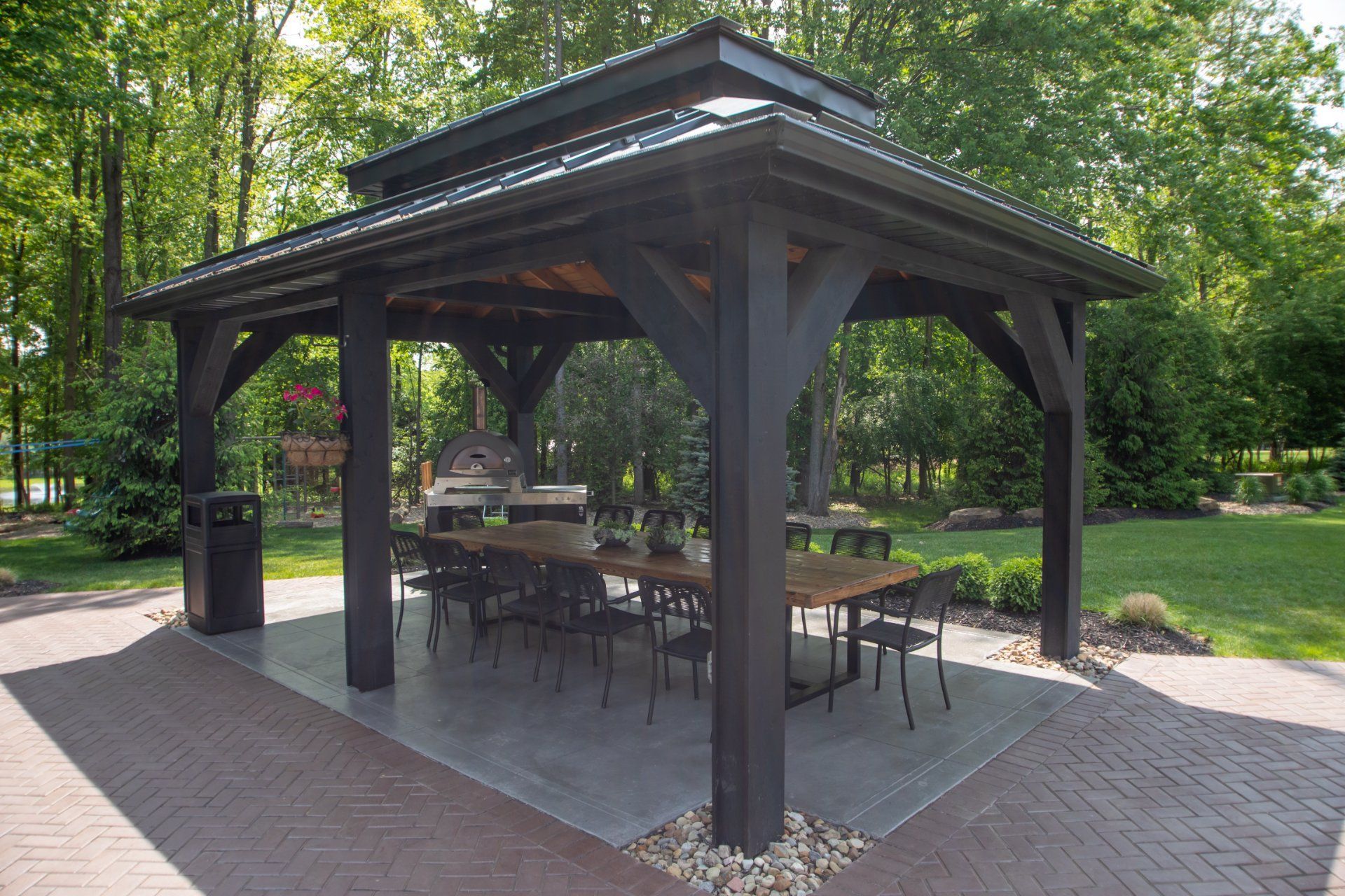 Black gazebo with a dining table and chairs on a patio with a grill, located in a park setting.
